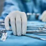 A gloved hand reaches for a surgical instrument on a table covered with various metal tools. The person is wearing blue surgical attire, following ANSI/AAMI ST108:2023 Standards, and the background is slightly out of focus, suggesting a medical or surgical setting.